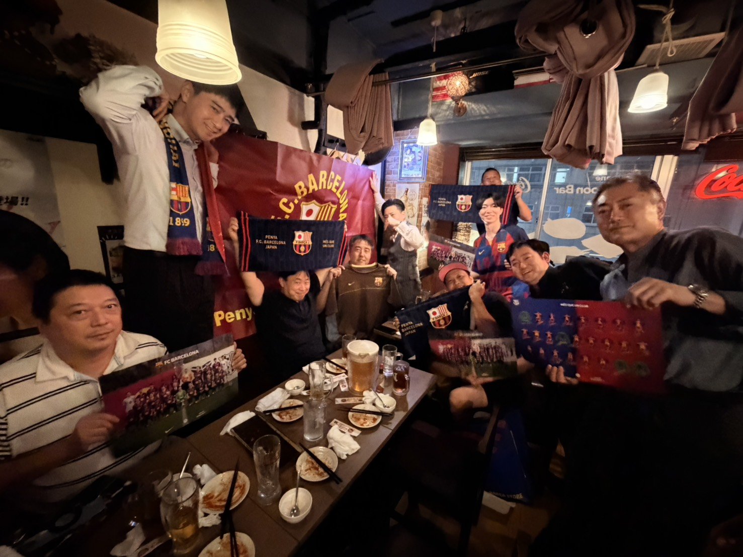 PBJ懇親会の集合写真。参加者がPBJのフラッグやマフラー、新グッズを手に笑顔で並んでいる